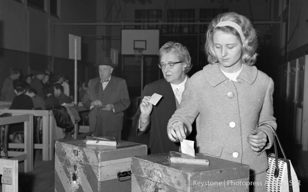 Photo en noir et blanc : Une femme plus âgée et une jeune femme lançant des bulletins de vote dans deux urnes